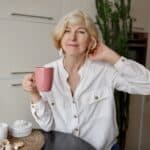 50-year-old blonde woman holding cup of tea and sitting at modern kitchen.