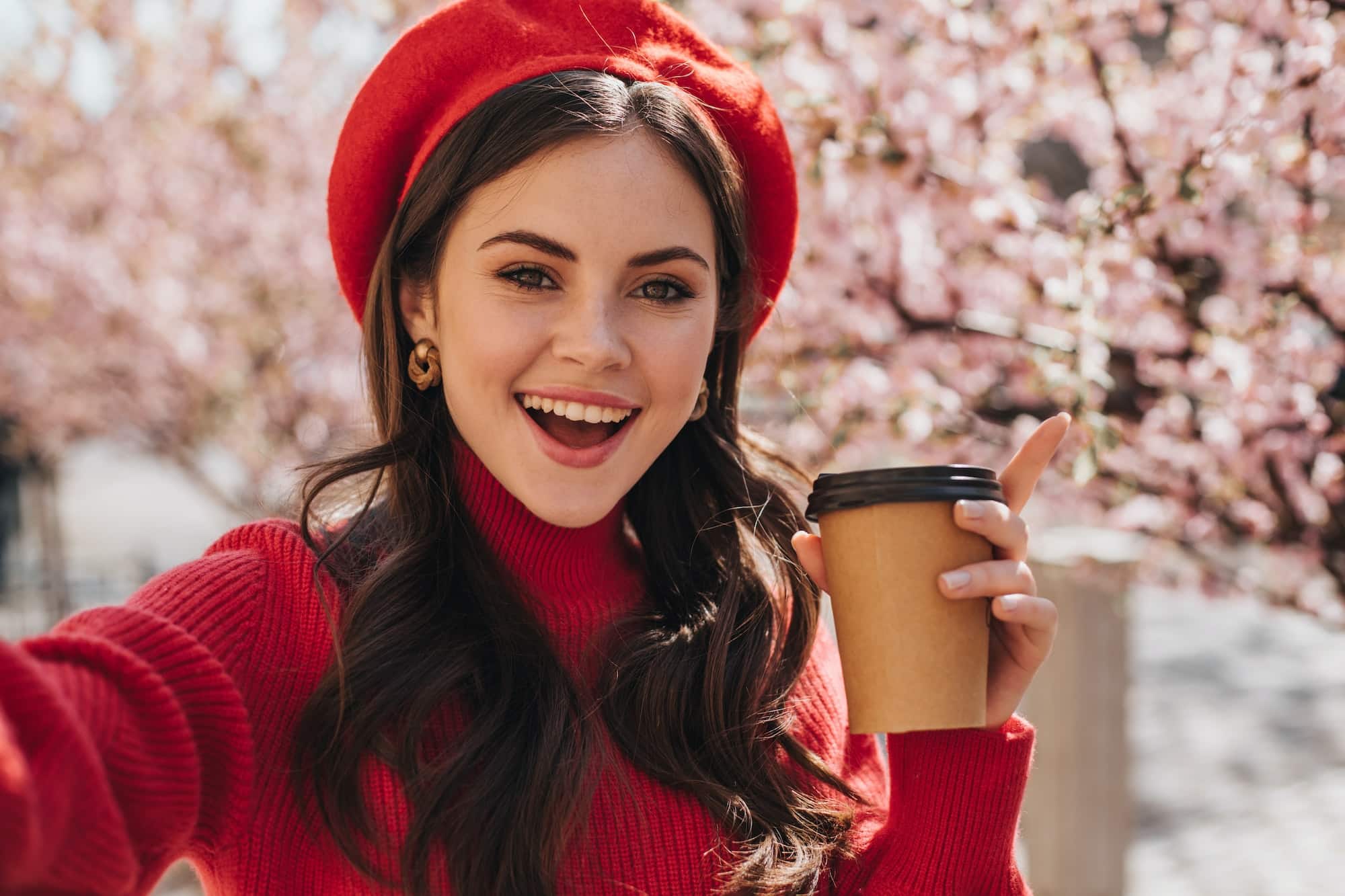 Beautiful woman in red outfit holds glass of tea and takes selfie on background of sakura. Portrait