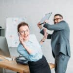 businessman get ready to hit his colleague with clipboard while she talking by phone and annoying