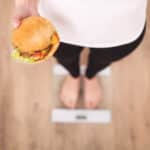 concept de régime et de restauration rapide. femme en surpoids debout sur une balance tenant un hamburger (hamburger). malbouffe malsaine. régime, mode de vie. perte de poids. obésité. vue de dessus