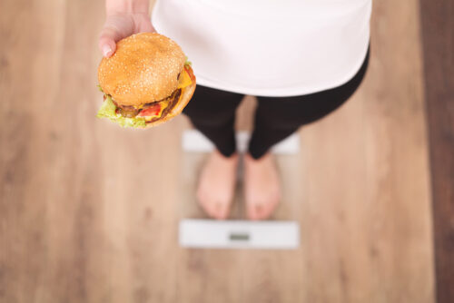 concept de régime et de restauration rapide. femme en surpoids debout sur une balance tenant un hamburger (hamburger). malbouffe malsaine. régime, mode de vie. perte de poids. obésité. vue de dessus