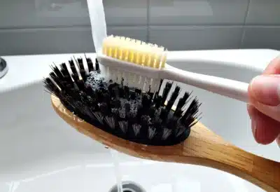 Main nettoyant une brosse à cheveux dans de l’eau tiède savonneuse avec une vieille brosse à dents pour retirer les résidus.